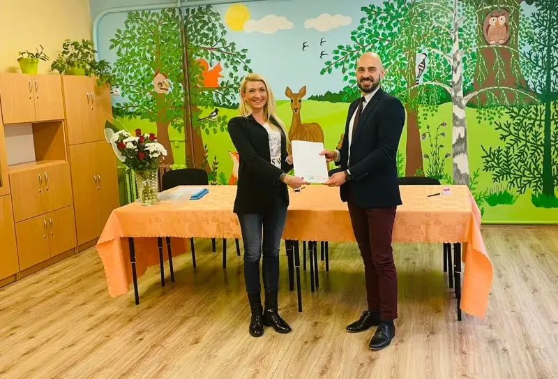 Zdjęcie o orientacji poziomej przedstawia dwie osoby stojące wewnątrz sali przedszkolnej, prawdopodobnie podczas uroczystego podpisania dokumentów lub przekazania umowy. Kobieta w czarnym żakiecie oraz mężczyzna w eleganckiej marynarce trzymają wspólnie białą kartkę papieru, stojąc przed długim stołem nakrytym brzoskwiniowym obrusem. Po lewej stronie stołu stoi szklany wazon z bukietem kwiatów, a w tle widać kolorowy, ręcznie malowany mural przedstawiający leśne zwierzęta, drzewa i słońce. Pomieszczenie posiada jasną podłogę z paneli oraz po lewej stronie wysokie, jasne szafki, co sugeruje przestrzeń edukacyjną dla dzieci. Oświetlenie wewnątrz jest równomierne i ciepłe, podkreślające formalny, a zarazem przyjazny charakter spotkania w placówce oświatowej.