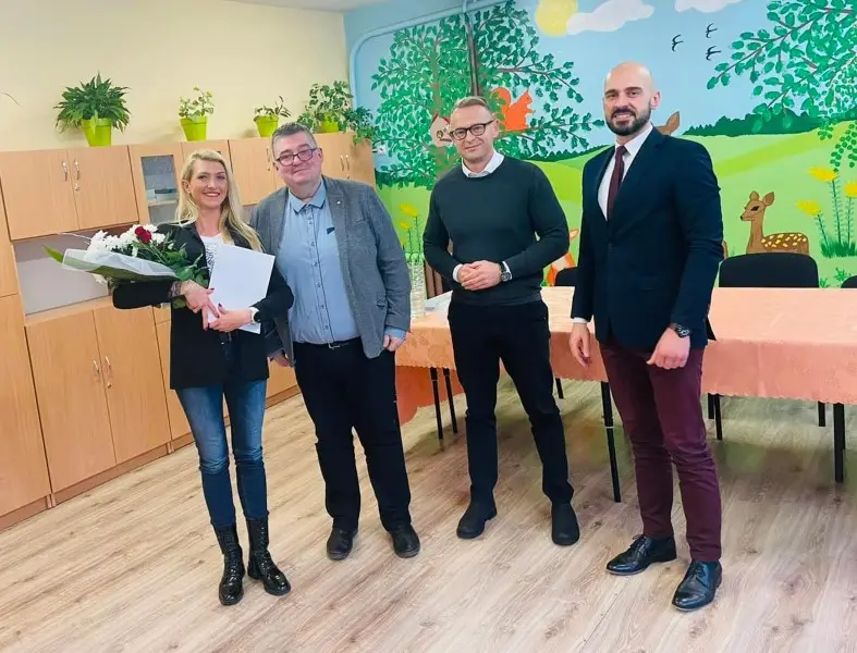 Zdjęcie wykonane w orientacji poziomej przedstawia grupę czterech osób stojących w jasnej sali przedszkolnej. Po lewej stronie znajduje się kobieta w czarnym żakiecie i jeansach, trzymająca bukiet kwiatów oraz białą teczkę. Obok niej stoją trzej mężczyźni: pierwszy w szarej marynarce i koszuli, drugi w ciemnozielonym swetrze, a trzeci w eleganckim garniturze z bordowym krawatem. W tle widać drewniane szafki, stół przykryty łososiowym obrusem oraz ścianę ozdobioną kolorowym malowidłem przedstawiającym leśne zwierzęta i drzewa. Cała scena jest dobrze oświetlona, co tworzy profesjonalną, a jednocześnie przyjazną atmosferę spotkania w placówce oświatowej.