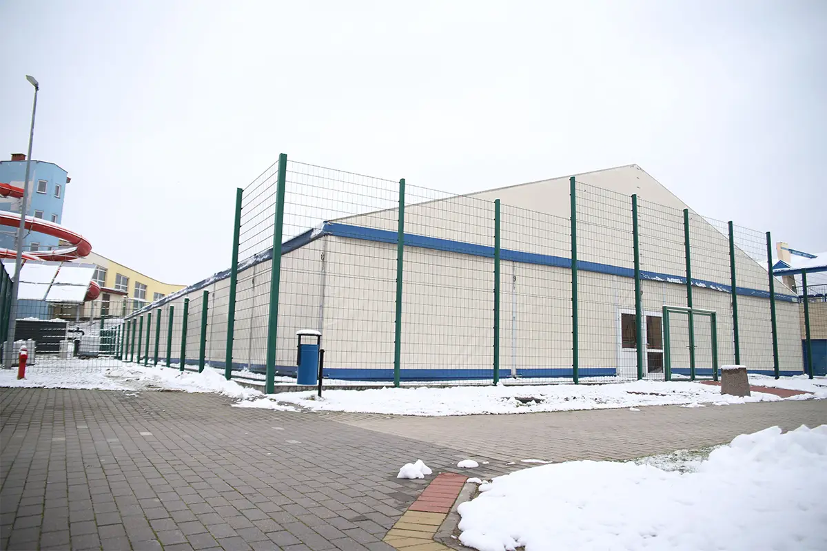 Zdjęcie przedstawia w układzie poziomym o proporcjach 3:2 nowoczesną halę sportową lub namiotową, która jest ogrodzona wysokim zielonym płotem z siatki stalowej. Jasne ściany obiektu z niebieskimi akcentami wyraźnie odcinają się od zaśnieżonego otoczenia, wskazując na zimową aurę. Na pierwszym planie widoczna jest wybrukowana kostką placówka z niewielkimi płatami śniegu oraz charakterystyczny niebieski kosz na śmieci. W lewej części kadru w tle wyłania się fragment niebieskiego budynku z czerwoną rurą zjeżdżalni, co sugeruje lokalizację w pobliżu obiektu rekreacyjnego lub aquaparku. Kompozycja zdjęcia jest szeroka, skupiając uwagę na bryle budynku oraz uporządkowanym zagospodarowaniu terenu wokół sportowej infrastruktury.